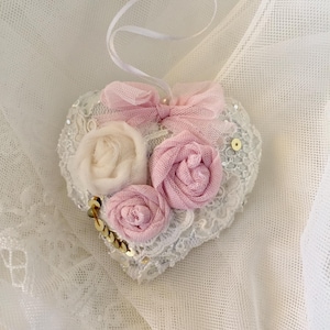 Op de afbeelding: Een wit hartvormig ornament met kantdetails en roze bloemen van stof. Het ornament is versierd met gouden pailletten.