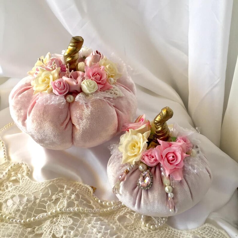 May include: Two pink velvet pumpkins decorated with lace, pearls, and pink and white flowers. The pumpkins are sitting on a white surface with a lace doily.