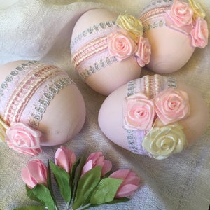 May include: Four pink Easter eggs decorated with lace and pink and yellow satin ribbons. The eggs are sitting on a white fabric surface.