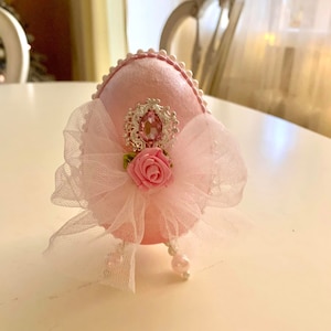 May include: A pink egg-shaped ornament decorated with a white tulle bow, a pink rose, and a silver and pink jeweled embellishment.