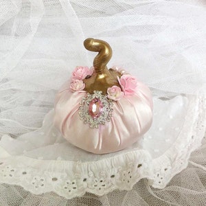 Könnte beinhalten: Ein rosa Satin-Kürbis mit goldenem Stiel und rosa Blumenakzenten. Ein Strassstein-Verzierung befindet sich mittig auf der Vorderseite des Kürbisses.