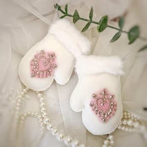May include: A pair of white felt mittens with pink and gold heart embellishments. The mittens are decorated with sparkling rhinestones and are perfect for adding a touch of whimsy to your festive decor.