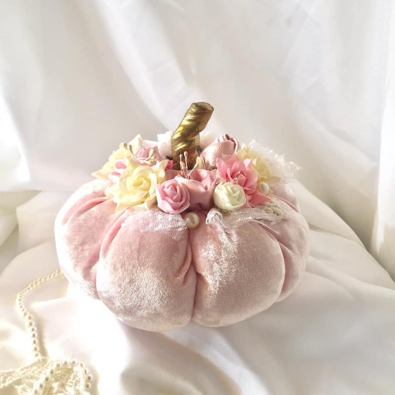 May include: A pink velvet pumpkin with a gold stem, decorated with pink, white and cream fabric flowers and lace.