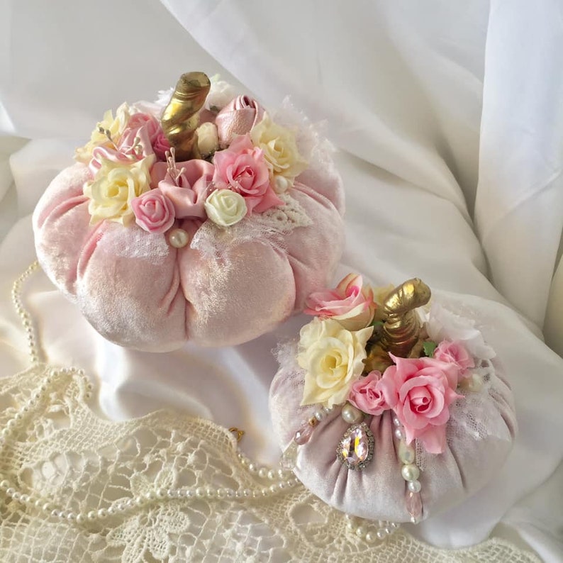 May include: Two pink velvet pumpkins decorated with lace, pearls, and pink and white flowers. The pumpkins are sitting on a white surface with a white lace doily.