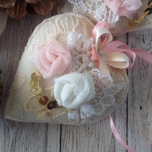 May include: A heart-shaped cushion with lace trim and embellishments. It features a pink and white rose, a pink ribbon, and gold sequins.