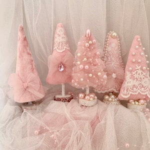 May include: Five pink Christmas trees decorated with lace, pearls, and tulle. The trees are arranged on a white tulle background.