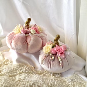 May include: Two pink velvet pumpkins decorated with lace, pearls, and pink and white flowers. The pumpkins are sitting on a white lace tablecloth.