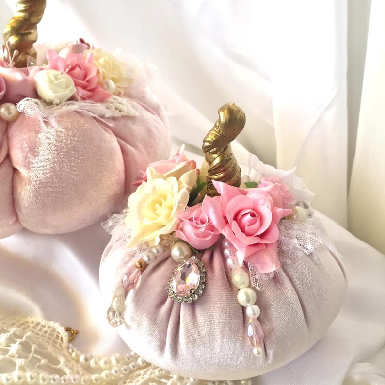 May include: Two pink velvet pumpkins decorated with white and pink fabric flowers, gold stems, pearls, and rhinestones.