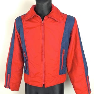 Può includere: Giacca rossa vintage con inserti blu navy sulle maniche e sui lati. La giacca presenta una cerniera a tutta lunghezza, due tasche con cerniera e cerniere sui polsi. Esposta su un manichino nero.