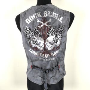 May include: Grey vest with a Rock Rebel design on the back. The graphic features two winged skulls, crossbones, and the text "Long Road Down, Life's a Gamble." The vest has a buckle closure and red stitching.