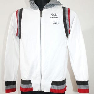 May include: White zip-up hooded jacket with gray accents and red trim. Features the text "G.S. G-star raw 3301" on the chest. The jacket has a ribbed hem and cuffs with gray, red, and black stripes.
