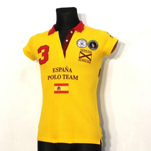 Puede incluir: Polo amarillo con un número 3 rojo, el texto "ESPAÑA POLO TEAM" y una pequeña bandera española en la parte delantera. La camiseta también tiene un cuello rojo y un parche con el texto "OFFICIAL SUPPLIER" en el lado derecho.