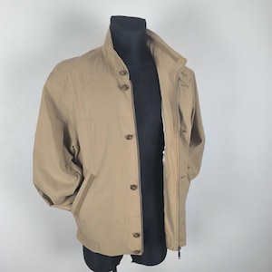 May include: A tan jacket with a high collar and button closure. The jacket has two front pockets and a zipper closure. The jacket is displayed on a black mannequin.