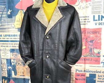 May include: A dark brown leather coat with a shearling collar and lining. The coat has a button-up front, two front pockets, and a yellow turtleneck sweater underneath. The coat is displayed on a black mannequin.