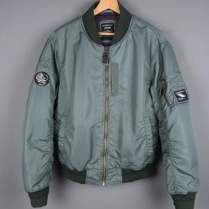 Puede incluir: Una chaqueta bomber verde salvia con cuello, puños y dobladillo verde oscuro. La chaqueta tiene cierre de cremallera, múltiples bolsillos y parches en las mangas. La chaqueta está colgada en una percha de madera.