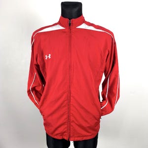 Puede incluir: Chaqueta deportiva roja con un acento blanco en los hombros y ribetes blancos. La chaqueta tiene una cremallera de largo completo y un pequeño logotipo blanco en el pecho. La chaqueta se exhibe en un maniquí negro.