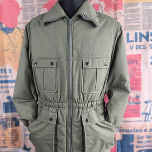 Puede incluir: Chaqueta verde oliva con cremallera, cuello y múltiples bolsillos. La chaqueta tiene cintura ceñida y detalles de botones. La chaqueta se muestra en una cabeza de maniquí negra. El fondo es un collage de recortes de periódicos.