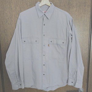 Puede incluir: Una camisa gris claro con botones, dos bolsillos en el pecho y mangas largas. La camisa tiene cuello y está abotonada por delante. Una pequeña etiqueta naranja es visible en uno de los bolsillos. La camisa está colgada en una percha de madera.