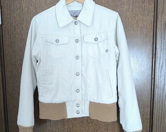 Puede incluir: Una chaqueta vaquera blanca con cuello y puños de pana marrón. La chaqueta tiene cierre de botones y dos bolsillos en la parte delantera.