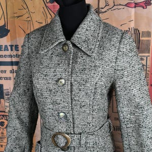 Puede incluir: Un abrigo de tweed gris con cuello con solapas, tres botones y un cinturón con una hebilla grande y redonda. El abrigo tiene un diseño clásico y está hecho de una tela texturizada.