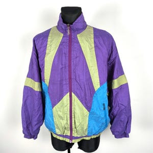 Puede incluir: Una chaqueta cortavientos de color morado, verde y azul con cierre de cremallera. La chaqueta tiene un diseño geométrico con paneles de colores contrastantes.