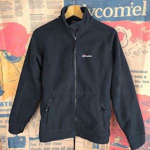 Puede incluir: Chaqueta polar negra con cremallera completa, dos bolsillos laterales y cuello alto. El logotipo de Berghaus está bordado en el pecho. La chaqueta está colgada en una percha de madera.