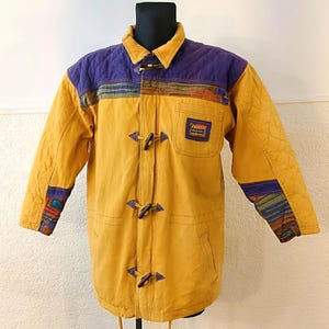 Puede incluir: Chaqueta de color amarillo mostaza con canesú y cuello morados acolchados. La chaqueta presenta cierres de palanca, un pequeño bolsillo y detalles estampados coloridos en las mangas y el pecho. La chaqueta se exhibe en un maniquí negro.