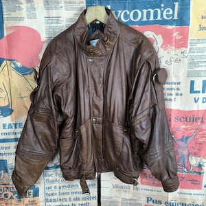 May include: A brown leather jacket with a high collar, multiple pockets, and silver-tone hardware. The jacket has a vintage style with a slightly worn appearance. The jacket is hanging on a wooden hanger.