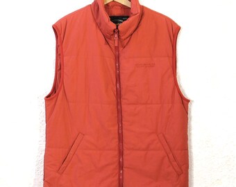 Puede incluir: Un chaleco acolchado de color naranja brillante con cuello alto, cremallera frontal completa y dos bolsillos laterales inclinados. La prenda presenta una construcción acolchada con costuras horizontales y un logotipo bordado sutil en el pecho.