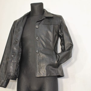 Puede incluir: Chaqueta de cuero negra con un diseño clásico, con solapas y cierre de botones. La chaqueta se exhibe en un maniquí negro, mostrando su ajuste a medida y su apariencia elegante. La chaqueta tiene un bolsillo a cada lado.