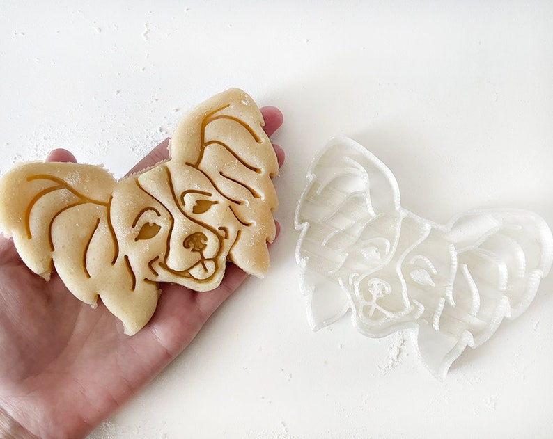 May include: A white plastic cookie cutter and a baked cookie in the shape of a Papillon dog's head.
