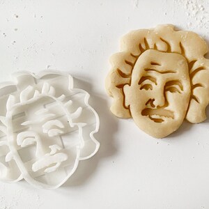 May include: A white plastic cookie cutter shaped like Albert Einstein's face, next to a baked cookie in the same shape.