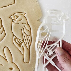 May include: A clear plastic cookie cutter shaped like a bird with a detailed design. The cutter is being held by a hand.