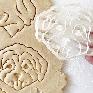 May include: A clear plastic dog-shaped cookie cutter being used to cut out a dog-shaped cookie from dough. The dough is light brown, and the cutter is held by a hand. The cookie cutter has a detailed design of a dog's face.