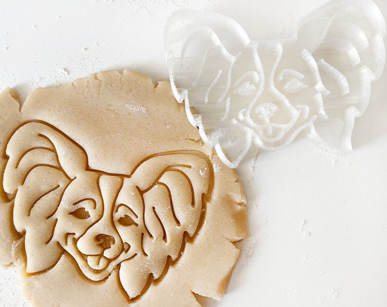 May include: A white plastic cookie cutter in the shape of a papillon dog is being used to cut out a cookie from a sheet of cookie dough.