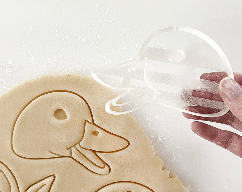 May include: A clear plastic cookie cutter shaped like a duck. The cutter is being held in a hand over a sheet of cookie dough.