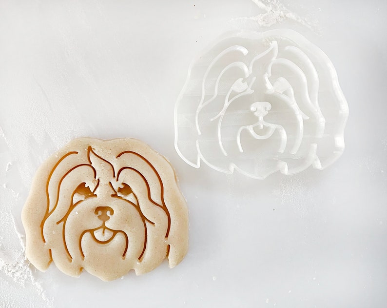 Puede incluir: Dos cortadores de galletas con forma de cabeza de perro. El cortador de la izquierda est&aacute; hecho de masa marr&oacute;n y el cortador de la derecha est&aacute; hecho de pl&aacute;stico transparente.