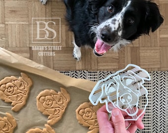 May include: A black and white dog looks up at the camera. A clear plastic dog-shaped cookie cutter is held in a hand. Several dog-shaped cookies are on a baking sheet. The text "Baker's Street Cutters" is visible.