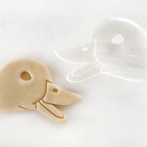May include: A close-up of a cookie cutter and a cookie in the shape of a duck's head. The cookie cutter is clear plastic and the cookie is a light brown color.