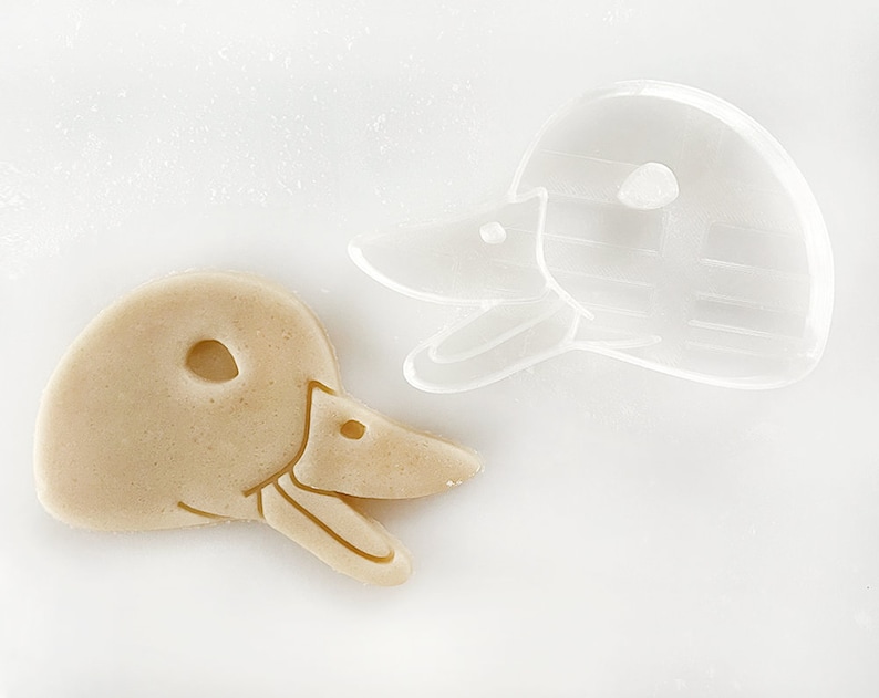 May include: A close-up of a cookie cutter and a cookie in the shape of a duck's head. The cookie cutter is clear plastic and the cookie is a light brown color.