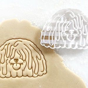 May include: A white plastic cookie cutter in the shape of a Komondor dog. The cutter is on top of a sheet of cookie dough, with the outline of the dog imprinted in the dough.