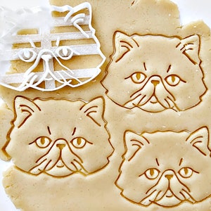 以下が含まれることがあります： ペルシャ猫の顔の形をしたクッキーカッター3つのクローズアップ。クッキーカッターは透明なプラスチック製で、クッキー生地のシートの上に置かれています。クッキー生地は薄い茶色で、クッキーカッターの形に押し込まれています。