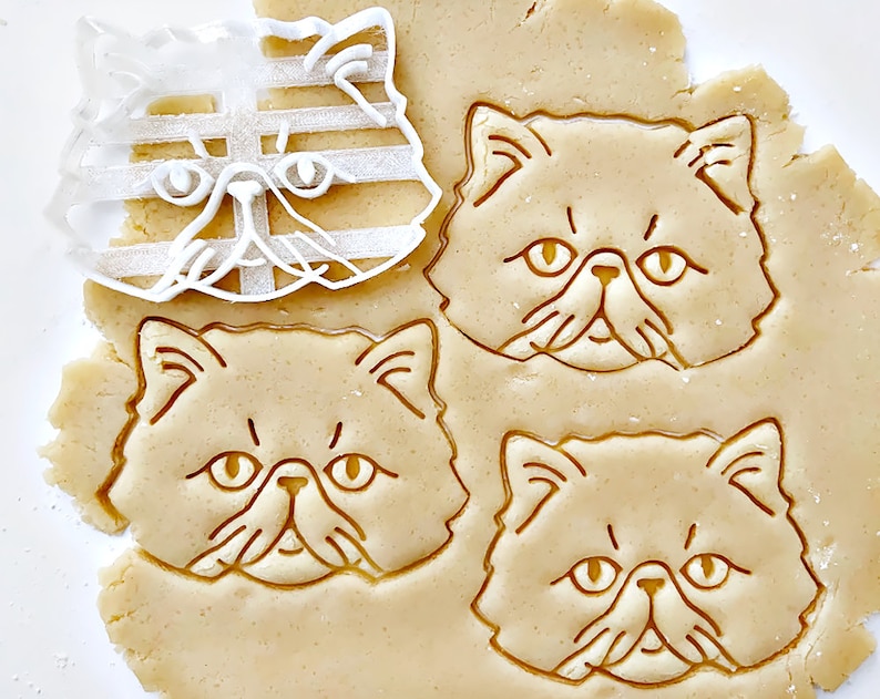以下が含まれることがあります： ペルシャ猫の顔の形をしたクッキーカッター3つのクローズアップ。クッキーカッターは透明なプラスチック製で、クッキー生地のシートの上に置かれています。クッキー生地は薄い茶色で、クッキーカッターの形に押し込まれています。
