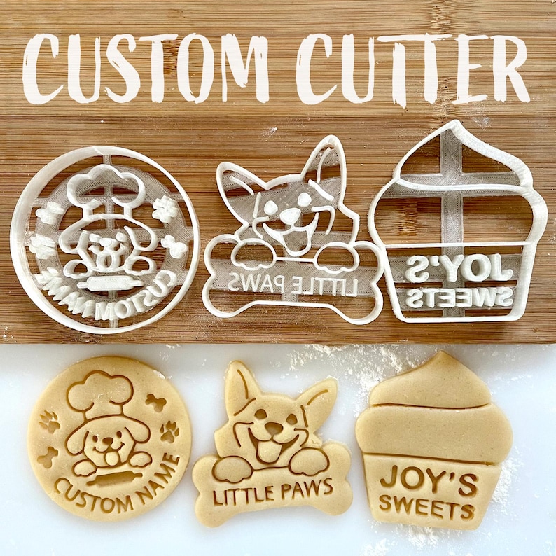 May include: A set of three cookie cutters, one shaped like a cupcake, one shaped like a dog with a bone, and one shaped like a chef with a hat. The cutters are made of white plastic and have the words "Joy's Sweets", "Little Paws", and "Custom Name" on them.