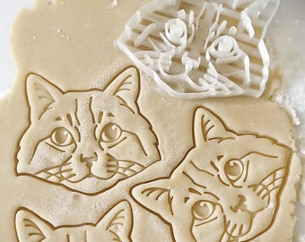 Puede incluir: Primer plano de tres cortadores de galletas con forma de gato. Los cortadores están hechos de plástico blanco y tienen un diseño detallado. Los cortadores están sobre una superficie marrón claro.