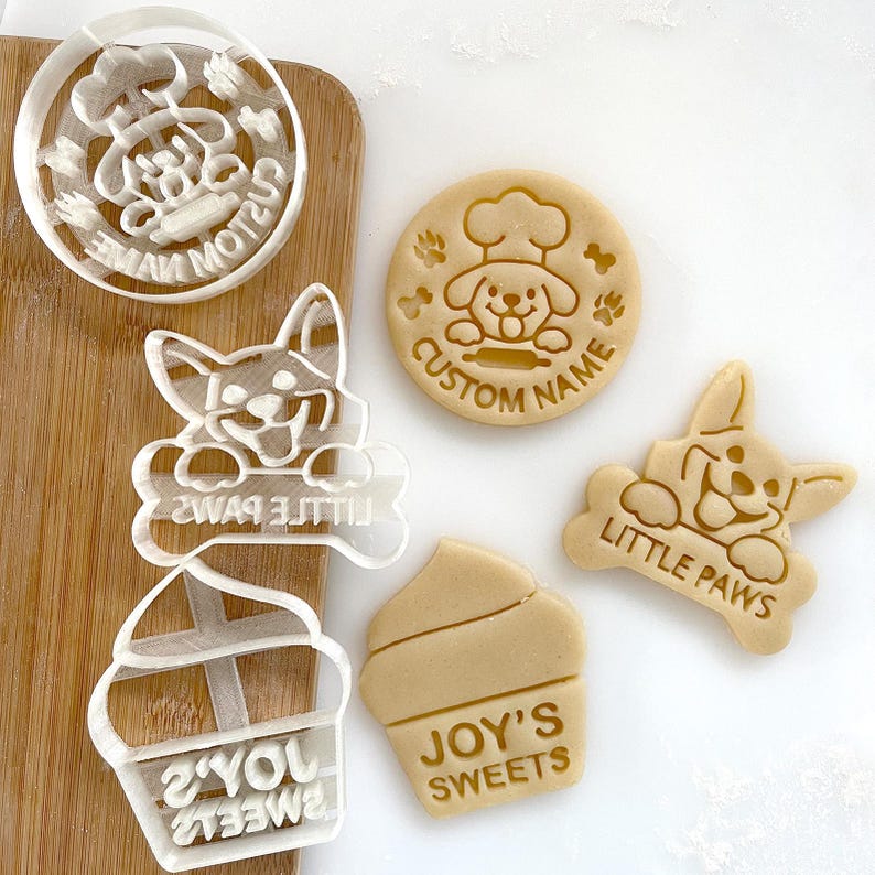 May include: A set of four cookie cutters featuring dog-themed designs. The cutters are made of clear plastic and feature various dog breeds, including a corgi, a chef dog, and a generic dog. The cutters are accompanied by three cookies, one with the text "Joy's Sweets", one with the text "Little Paws", and one with the text "Custom Name".