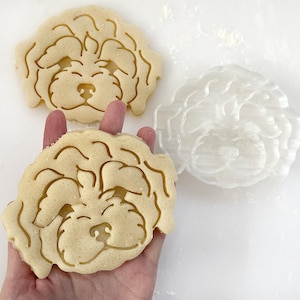 May include: Two sugar cookie cutouts in the shape of a dog's head. The cookies are light brown and have a detailed design. One cookie is being held in a hand.