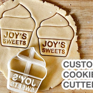 May include: A white plastic cookie cutter in the shape of a cupcake with the text "JOY'S SWEETS" on the front. The cookie cutter is on top of a piece of cookie dough with two other cupcake shapes already cut out. The text "CUSTOM COOKIE CUTTER" is written in grey below the cookie cutter.