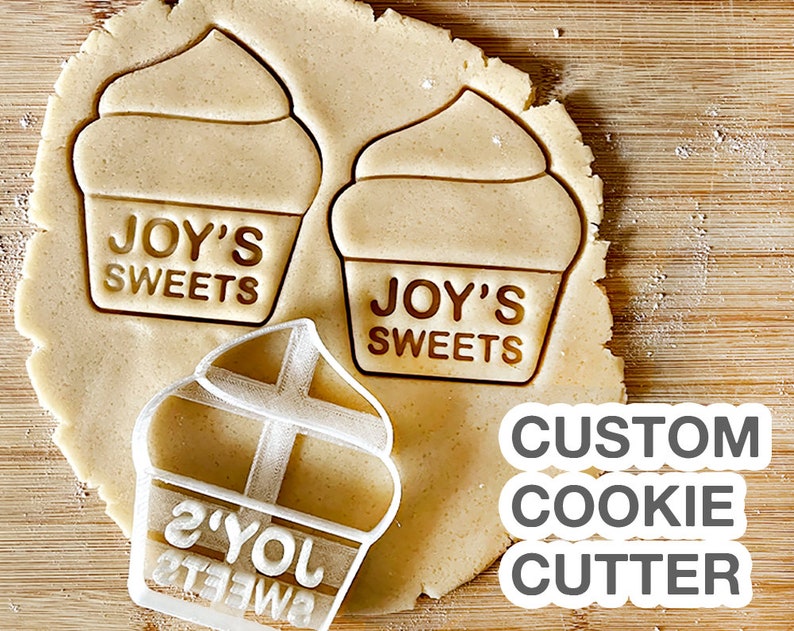 May include: A white plastic cookie cutter in the shape of a cupcake with the text "JOY'S SWEETS" on the front. The cookie cutter is on top of a piece of cookie dough with two other cupcake shapes already cut out. The text "CUSTOM COOKIE CUTTER" is written in grey below the cookie cutter.