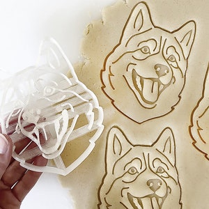 以下が含まれることがあります： ハスキー犬の顔の形をしたクッキー型3つのクローズアップ。クッキー型は透明なプラスチック製で、クッキー生地のシートの上に置かれています。